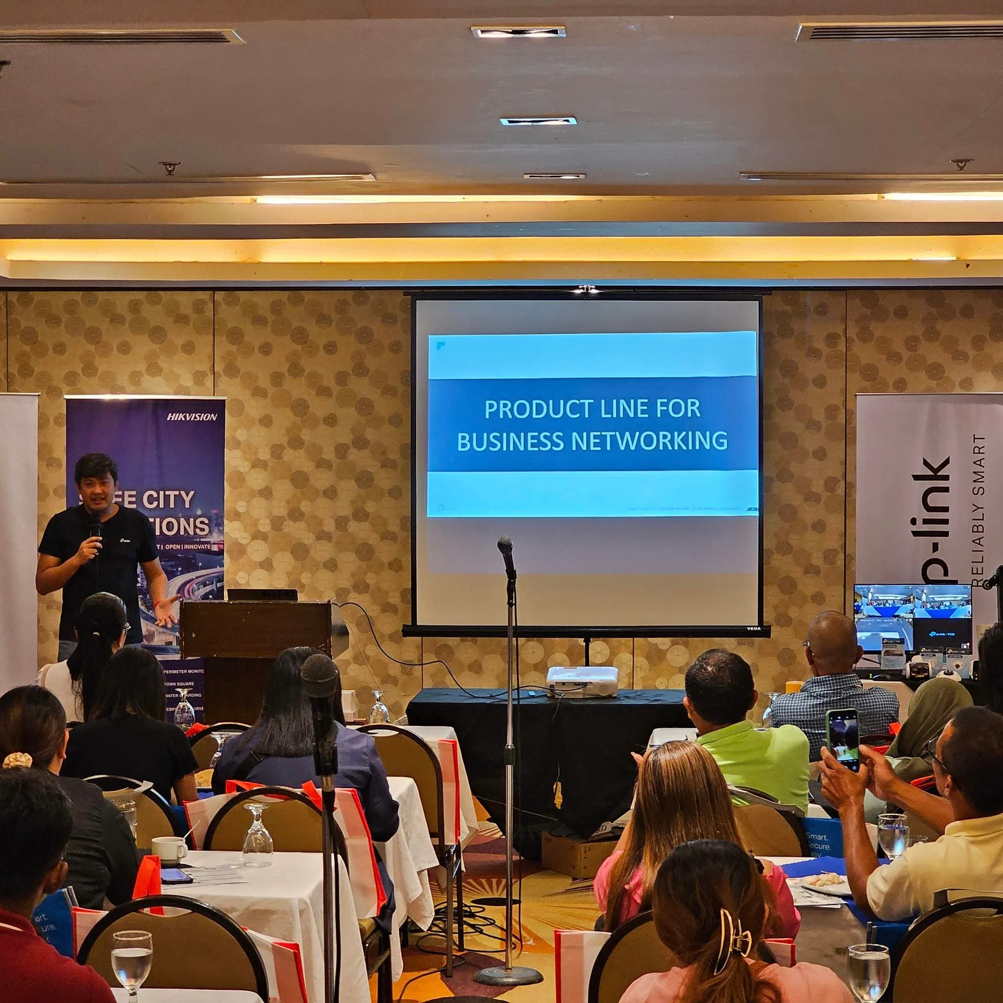 A speaker presents on stage at a conference, addressing an audience seated at tables. A screen displays "Product Line for Business Networking."