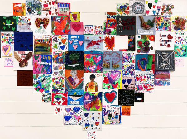 A concrete block wall is decorated with dozens of pieces of colorful student artwork arranged in the shape of a giant heart.