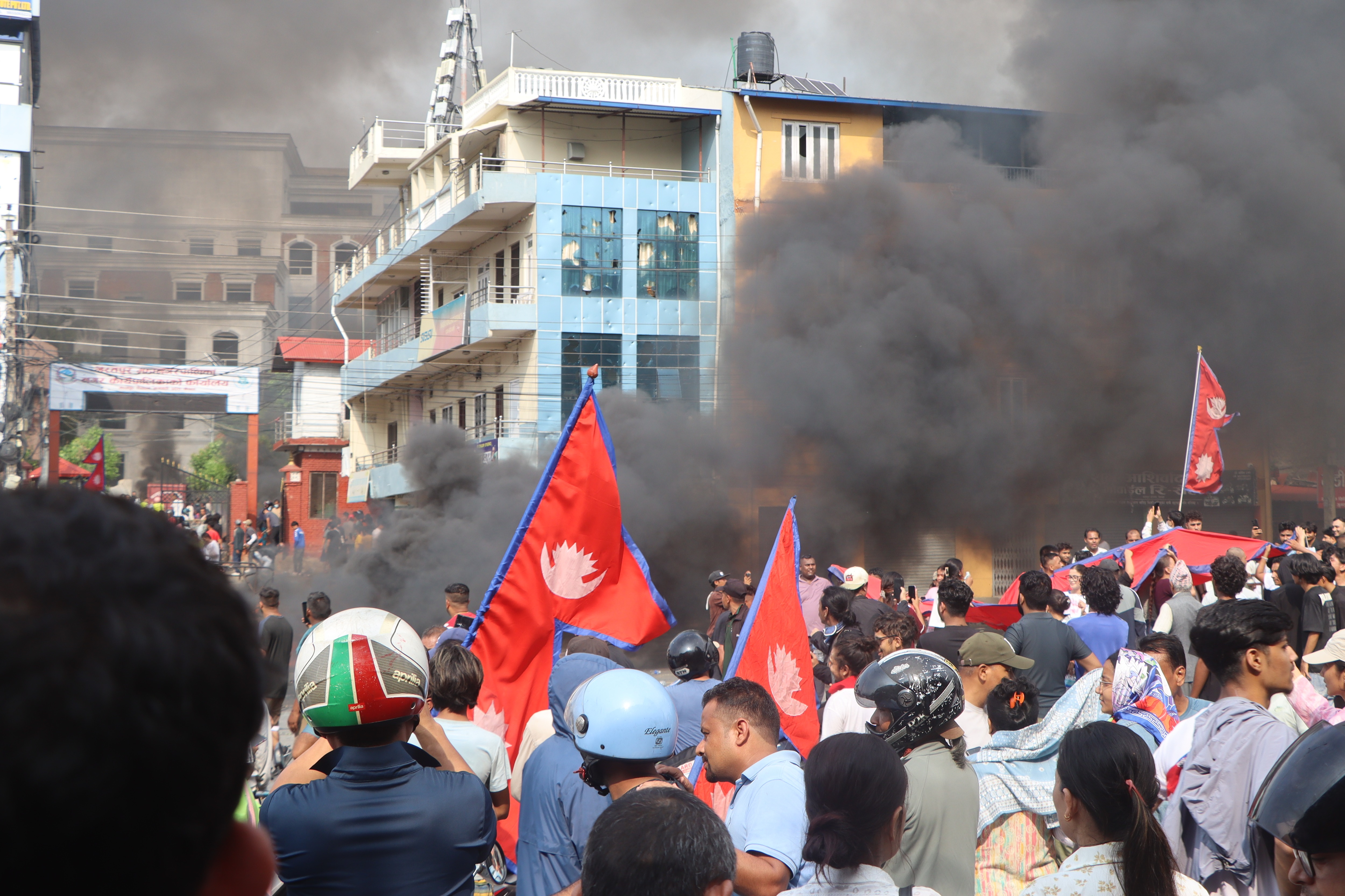 A large crowd of protesters carrying Nepalese flags stands in front of smoke-filled streets and burning buildings in an urban area, with thick black smoke rising behind them.