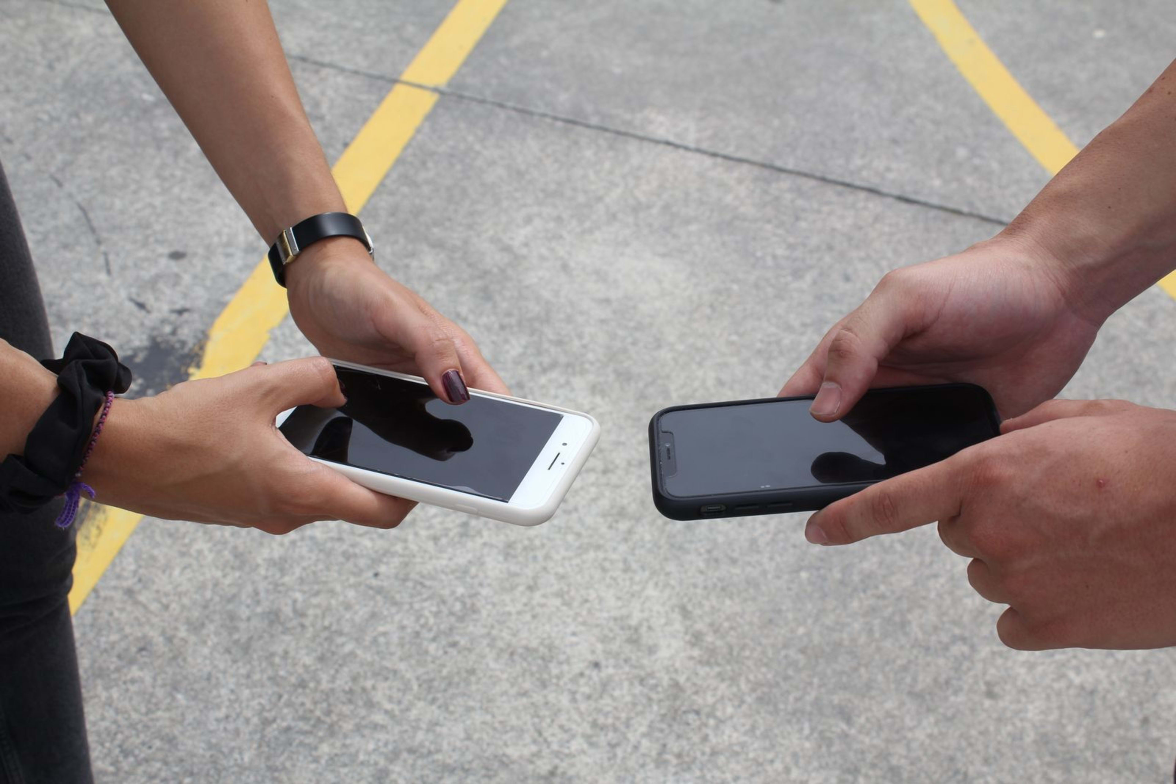 Two people holding smartphones facing each other outdoors, as if comparing the two phones side by side.