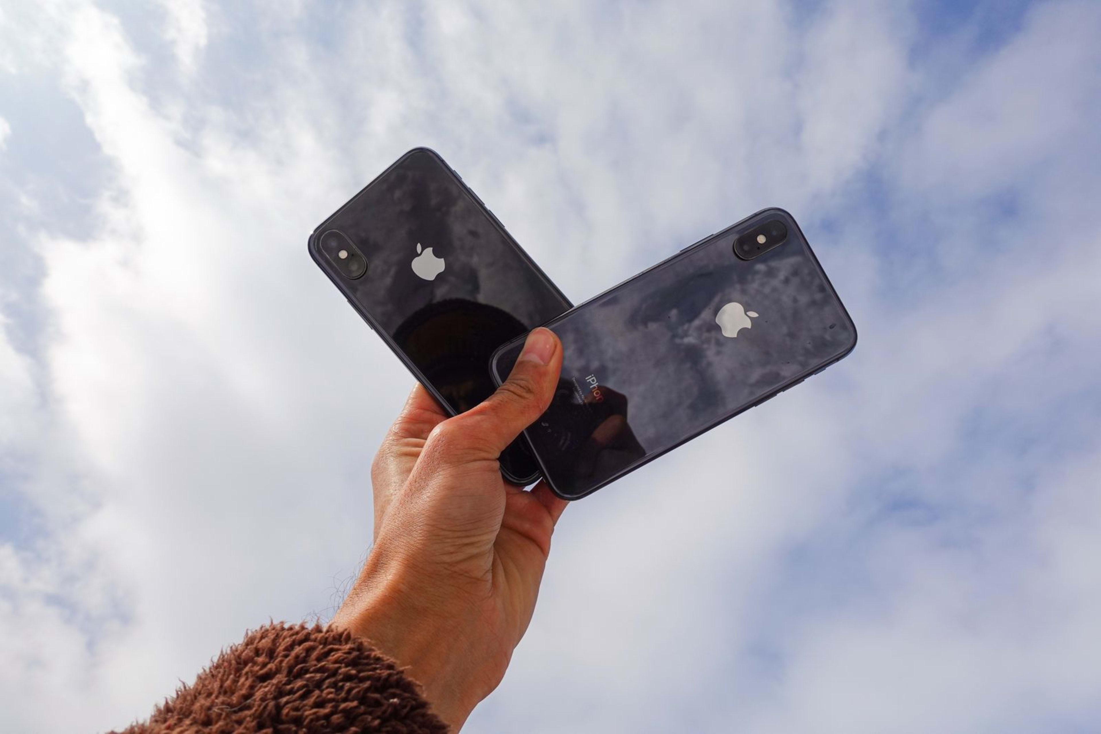 A hand holds up two black smartphones against a cloudy sky.