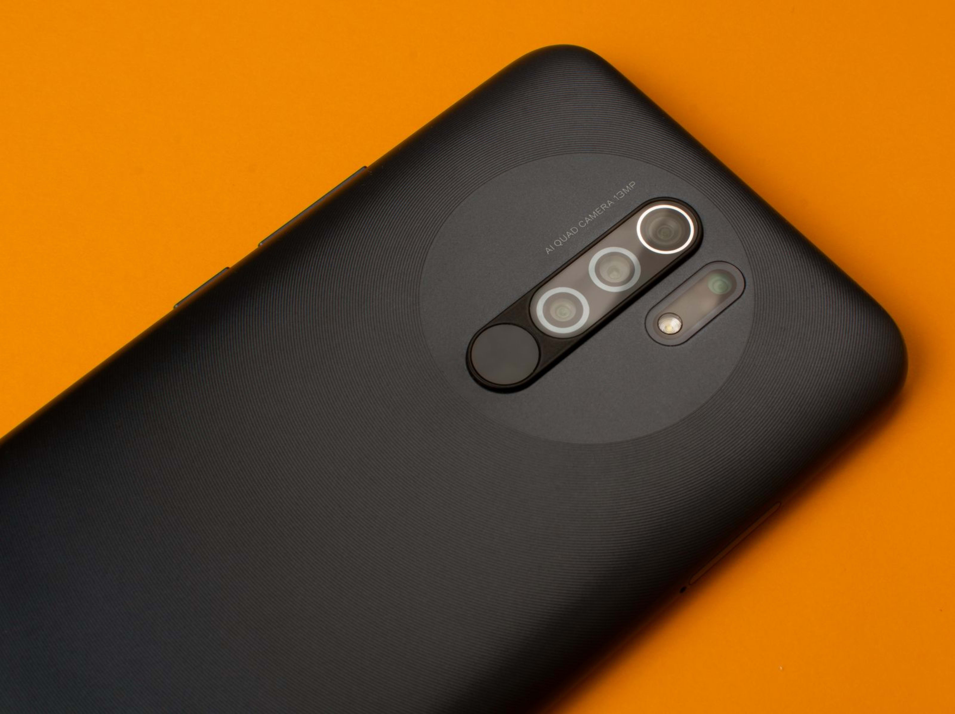 A close-up of a black smartphone's rear quad camera module on a bright orange background.