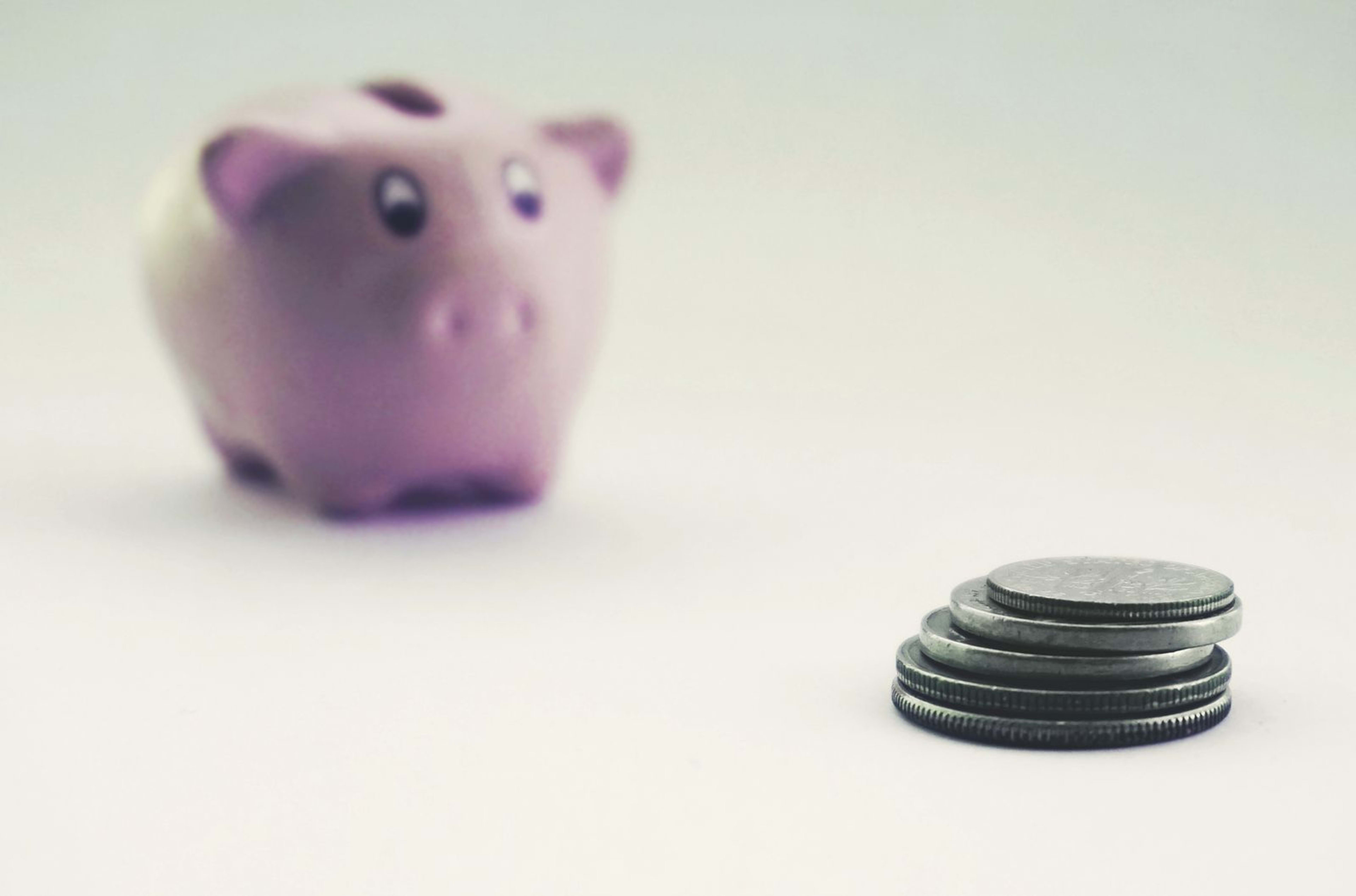 A pink piggy bank blurred in the background with a stack of coins in sharp focus on a white surface.