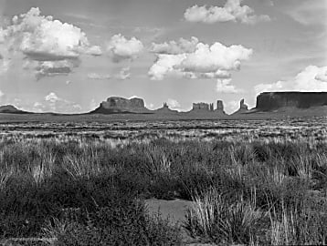 Black and white landscape photograph of Monument Valley with grassy foreground and puffy white clouds