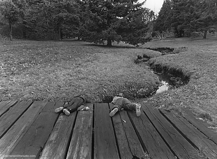 Black and white photograph of a foot bridge, stream, trees and two children lying on the bridge