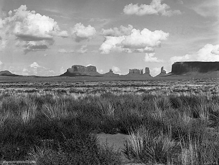Black and white landscape photograph of Monument Valley with grassy foreground and puffy white clouds
