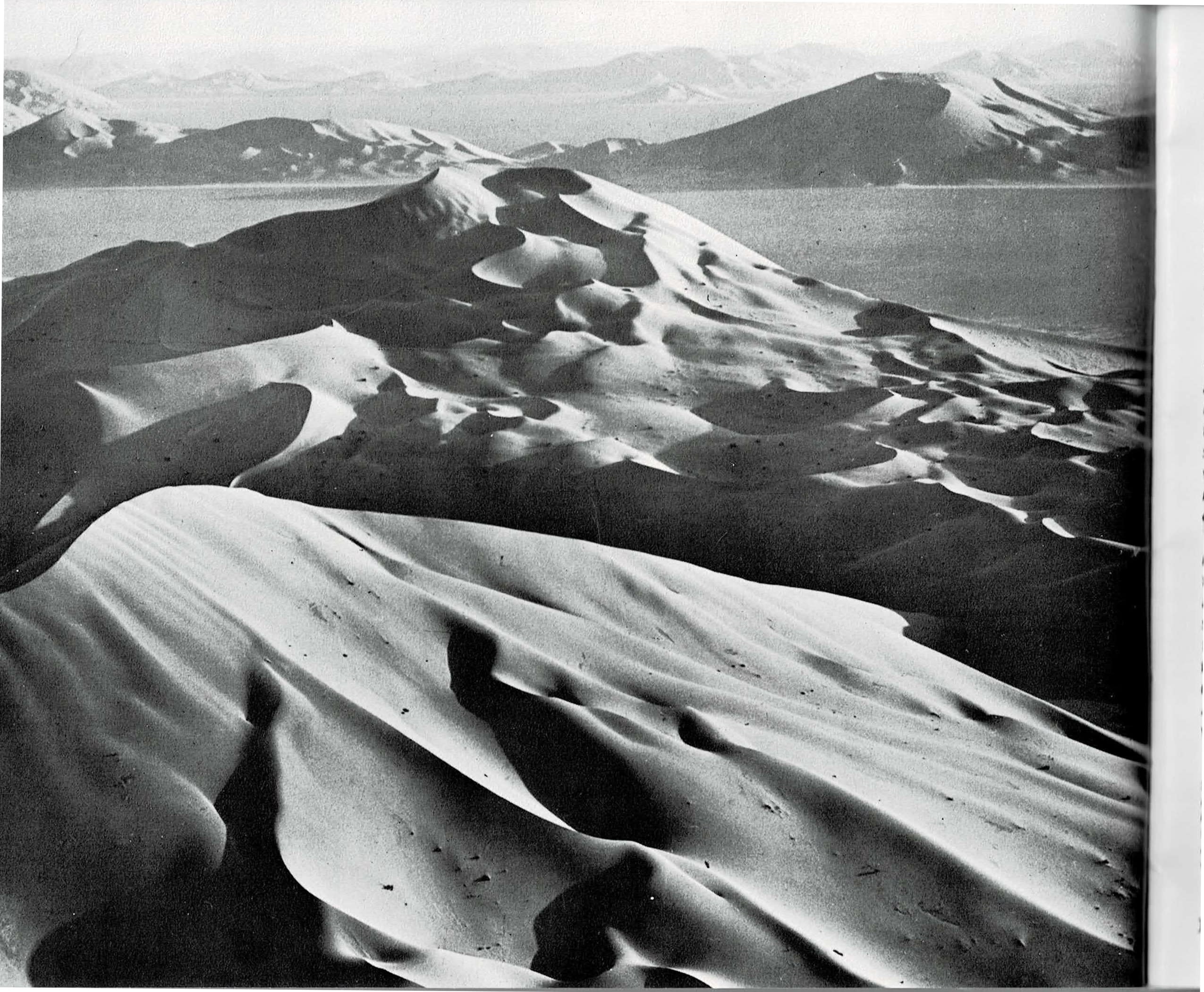 A two-page spread: at left, a full-bleed black and white aerial photograph of towering sand dunes in Saudi Arabia's Empty Quarter, their crests lit and rippled against a hazy desert horizon; at right, a text column recounting the 1872 Powell Survey's visit to Zion and quoting geologist Clarence Dutton's description of the Temples and Towers of the Virgin, beside a black and white photograph of dark, water-carved potholes and narrow sandstone walls.