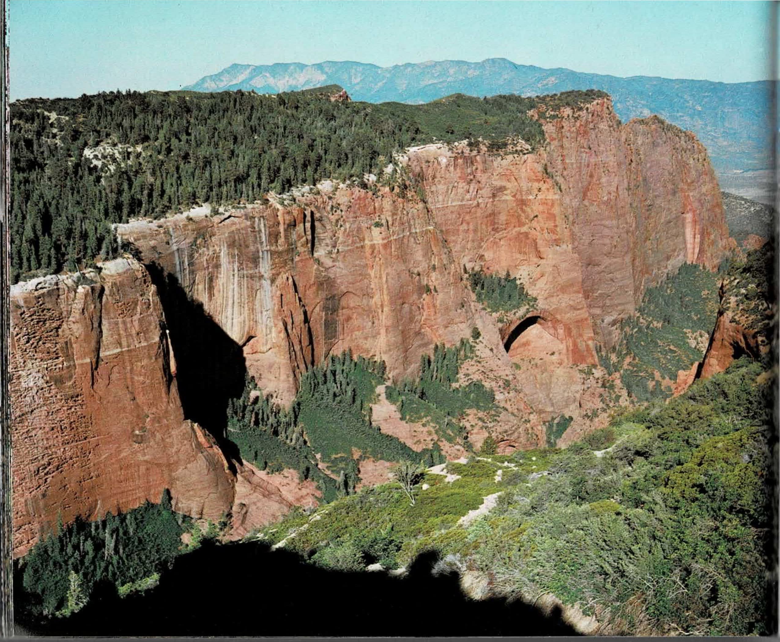 A two-page spread: at left, a full-bleed color photograph of a red-rock finger canyon in the Kolob, cliffs rising above forested slopes with a natural arch or alcove visible partway up the rock face and blue-hazed mountains receding into the distance; at right, a text column continuing mid-sentence, paired with a black and white photograph of a steep, crossbedded sandstone canyon dusted with snow and framed by conifers along its rim.