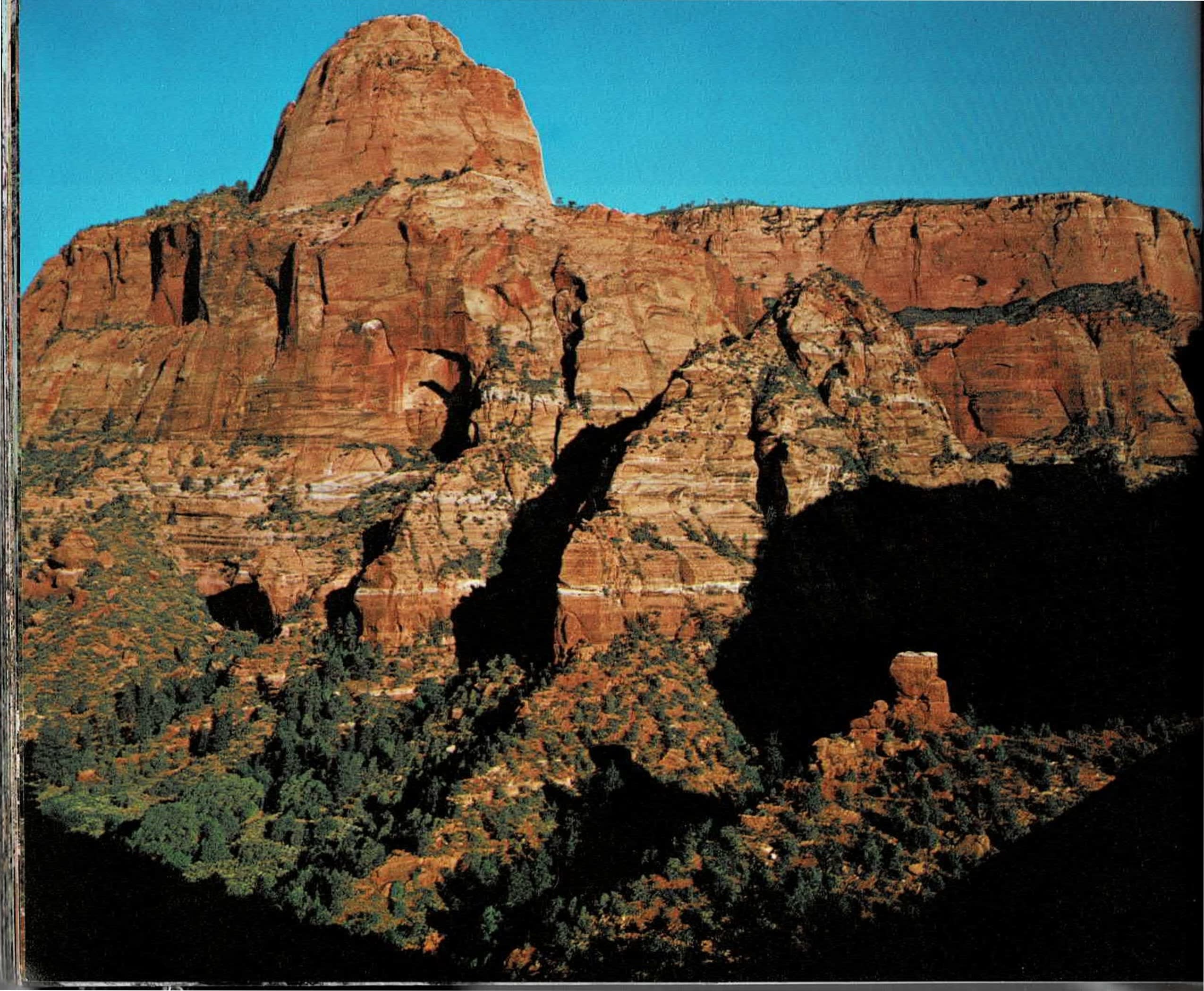 Color photograph of a sunlit sandstone butte with steep vertical cliffs and juniper-covered slopes on the left page, facing a printed text column and an aerial color photograph of a paved canyon road curving through a red rock canyon bend on the right page.