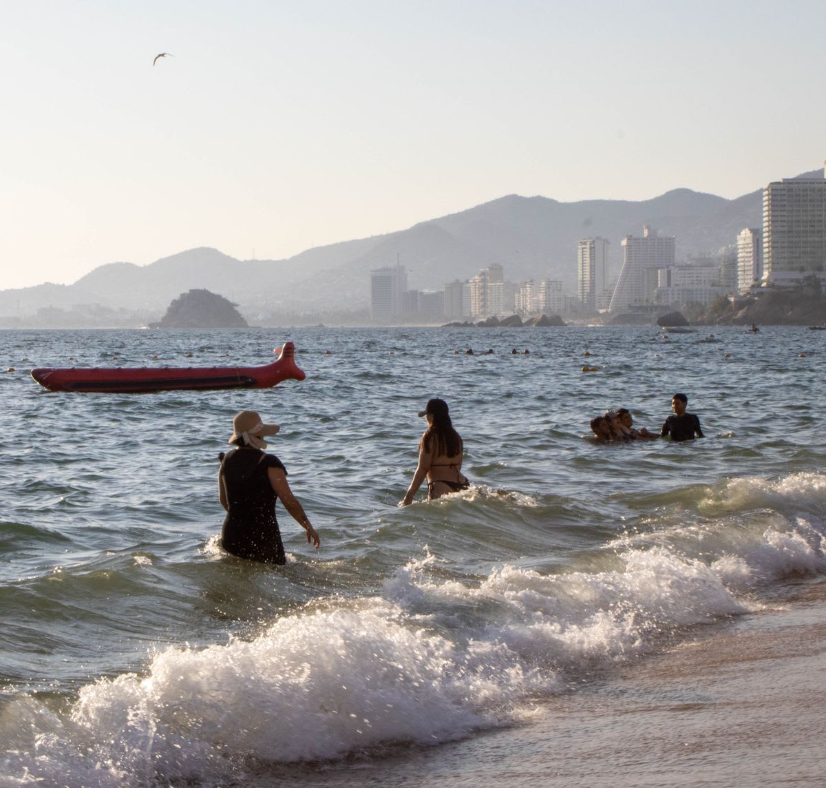 People enjoying the waves and beach at Acapulco with hotels visible in the background