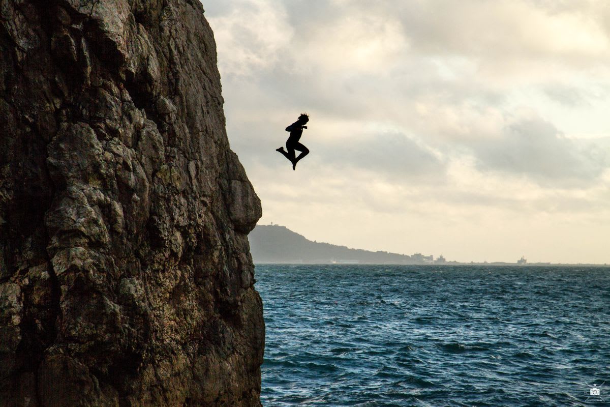 Person leaping from a rocky cliff into turquoise ocean water far below