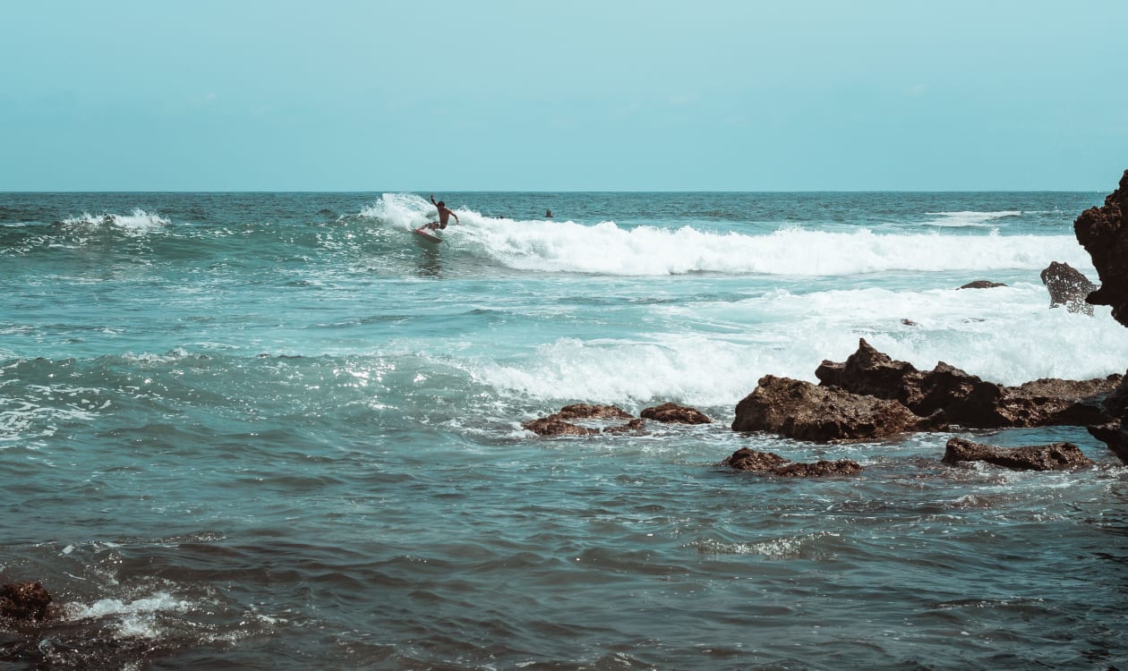 Surfer riding a wave along Puerto Escondido's rocky coastline in October, with clear skies overhead