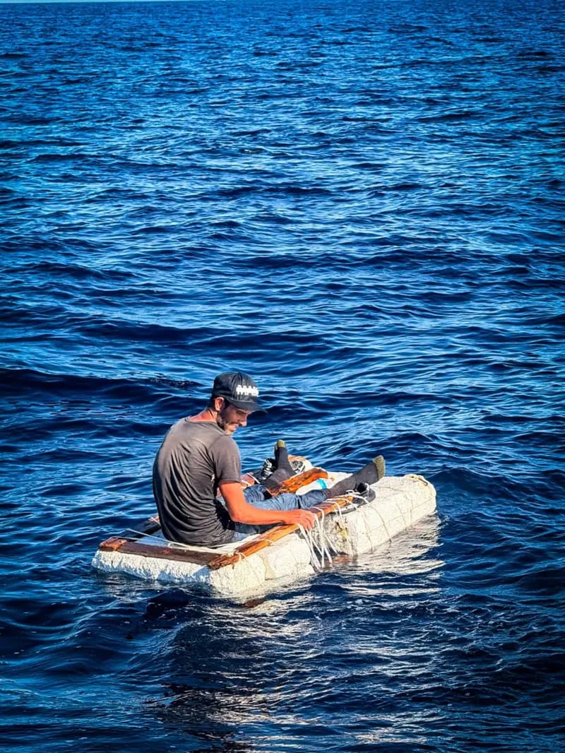Un hombre sentado sobre una balsa improvisada que flota en el mar.