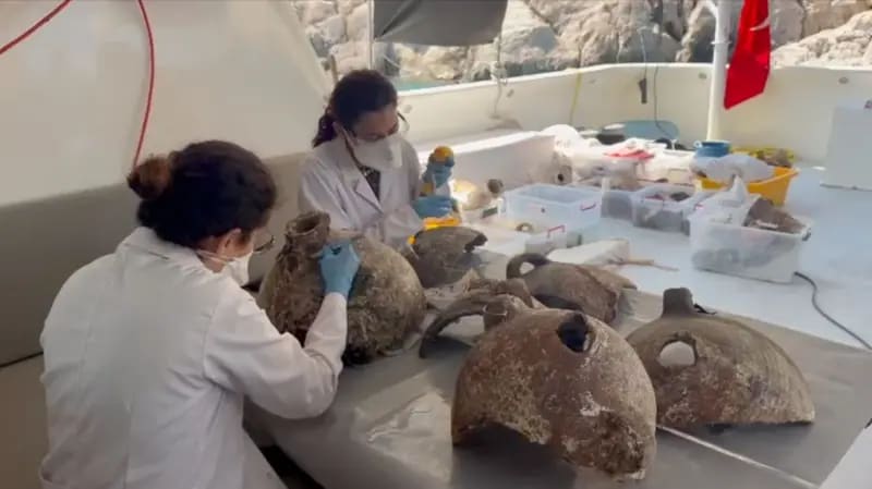 Dos personas con batas de laboratorio, guantes azules y mascarillas examinan algunas de las ánforas recuperadas. Están sentadas a una mesa en la parte trasera de un barco de buceo, a la sombra del tec