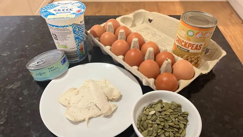 La foto muestra una lata de atún, un plato de pollo cocido, un tazón de semillas de calabaza, un envase de yogur griego, una caja de huevos y una lata de garbanzos sobre una encimera de cocina negra.