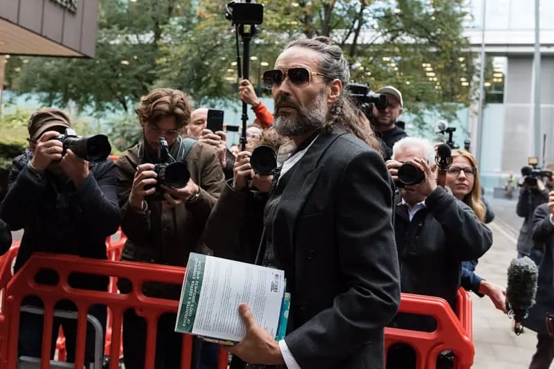 Russell Brand arriba al tribunal en Londres, con documentos en una mano. Varios fotógrafos lo enfocan al fondo.