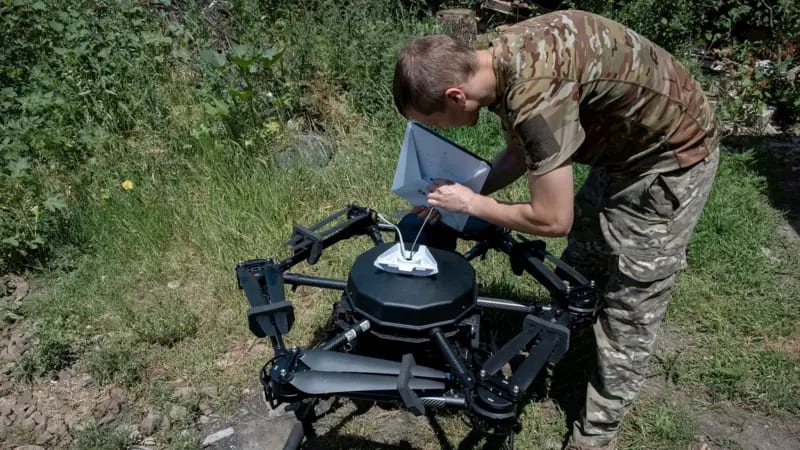 Un soldado con uniforme militar conecta un dispositivo de internet satelital a un gran dron negro en medio de una zona de matorrales verdes.