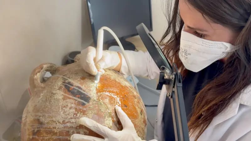 Primer plano de una ánfora en el laboratorio del barco. El asa del recipiente sobresale hacia la izquierda. Una mujer de pelo largo castaño, con bata blanca y mascarilla, trabaja en la superficie de l