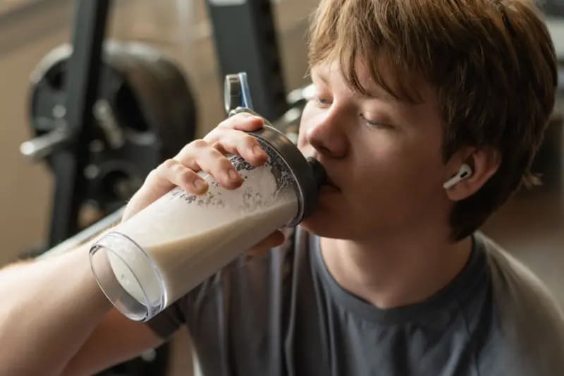 Un joven en un gimnasio toma un batido de proteína. Viste una camiseta deportiva gris, tiene audífonos y en el fondo se ven unas pesas