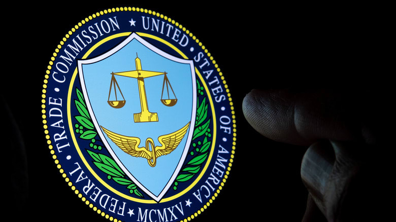 Photo of a black screen displaying the insignia for the US Federal Trade Commission. Two fingers near the image are faintly illuminated by the ambient light.