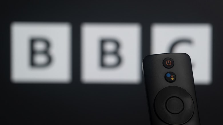 Photo of a smart tv remote held in front of a blurred BBC logo on a tv screen.