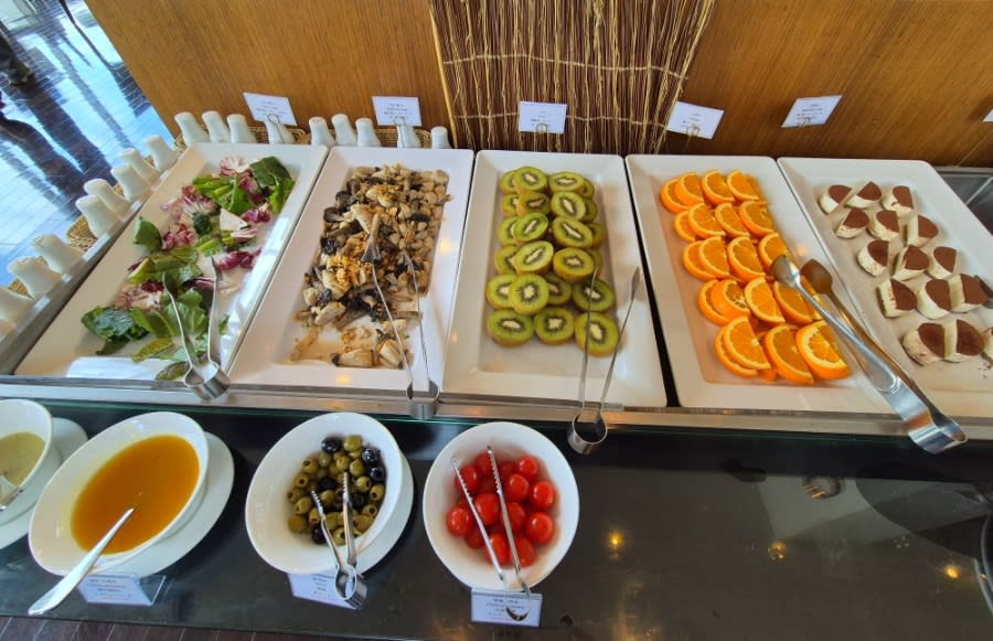 Buffet trays of salad, sliced kiwi, sliced oranges and small desserts, with bowls of sauce, olives and cherry tomatoes in front