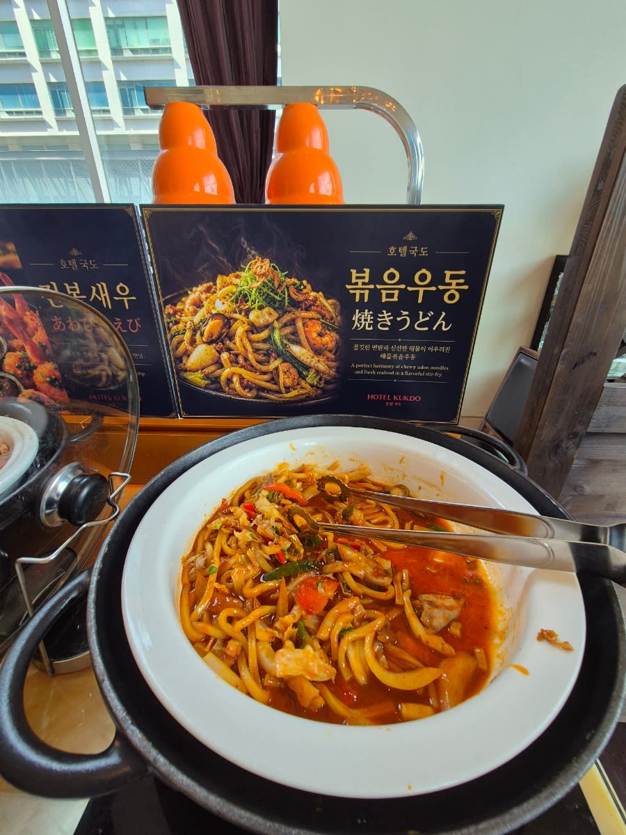 Hot buffet station with a white bowl of stir-fried udon and tongs