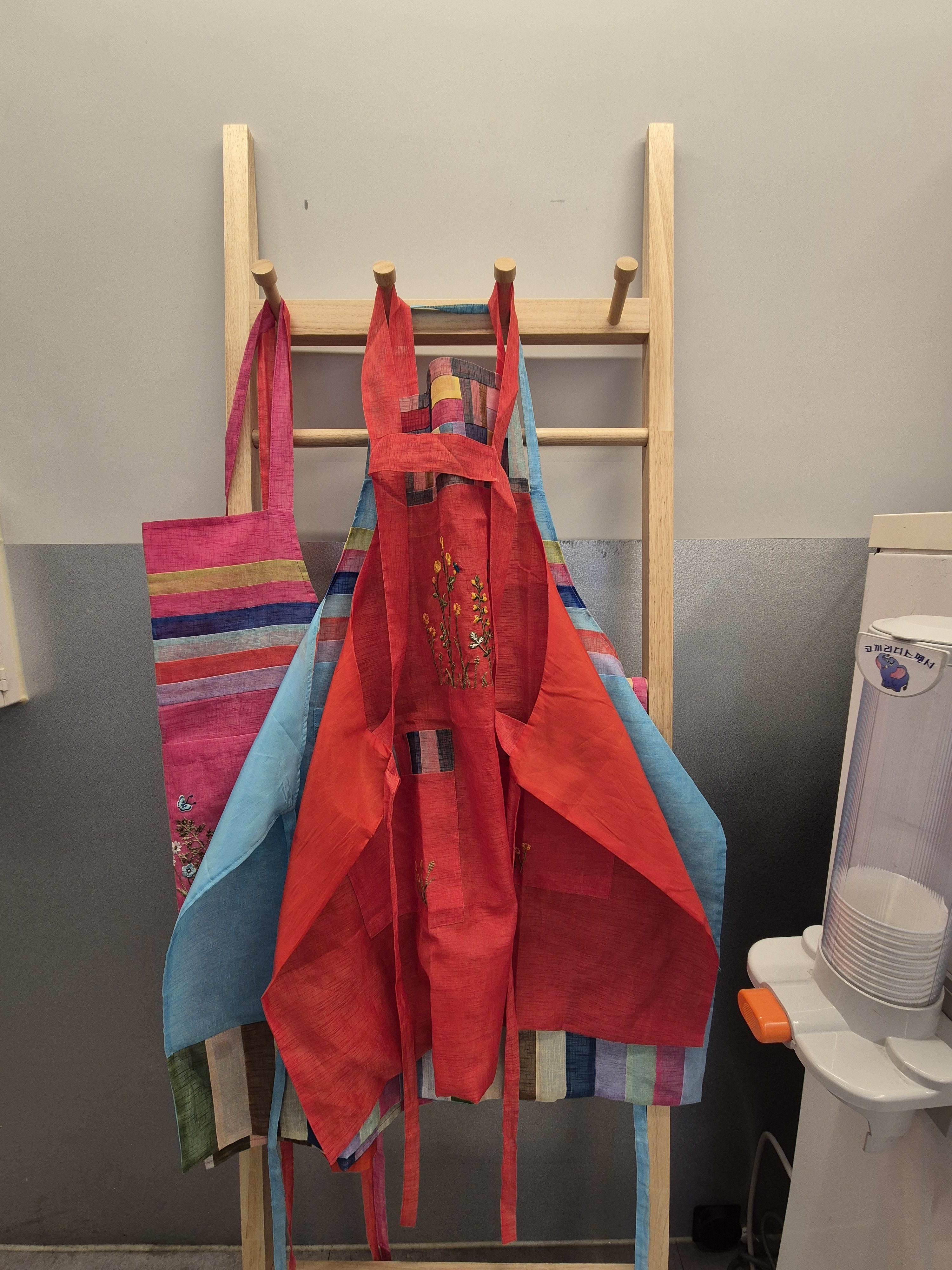 Two hanbok-style aprons hanging on a wooden rack, one red with floral embroidery