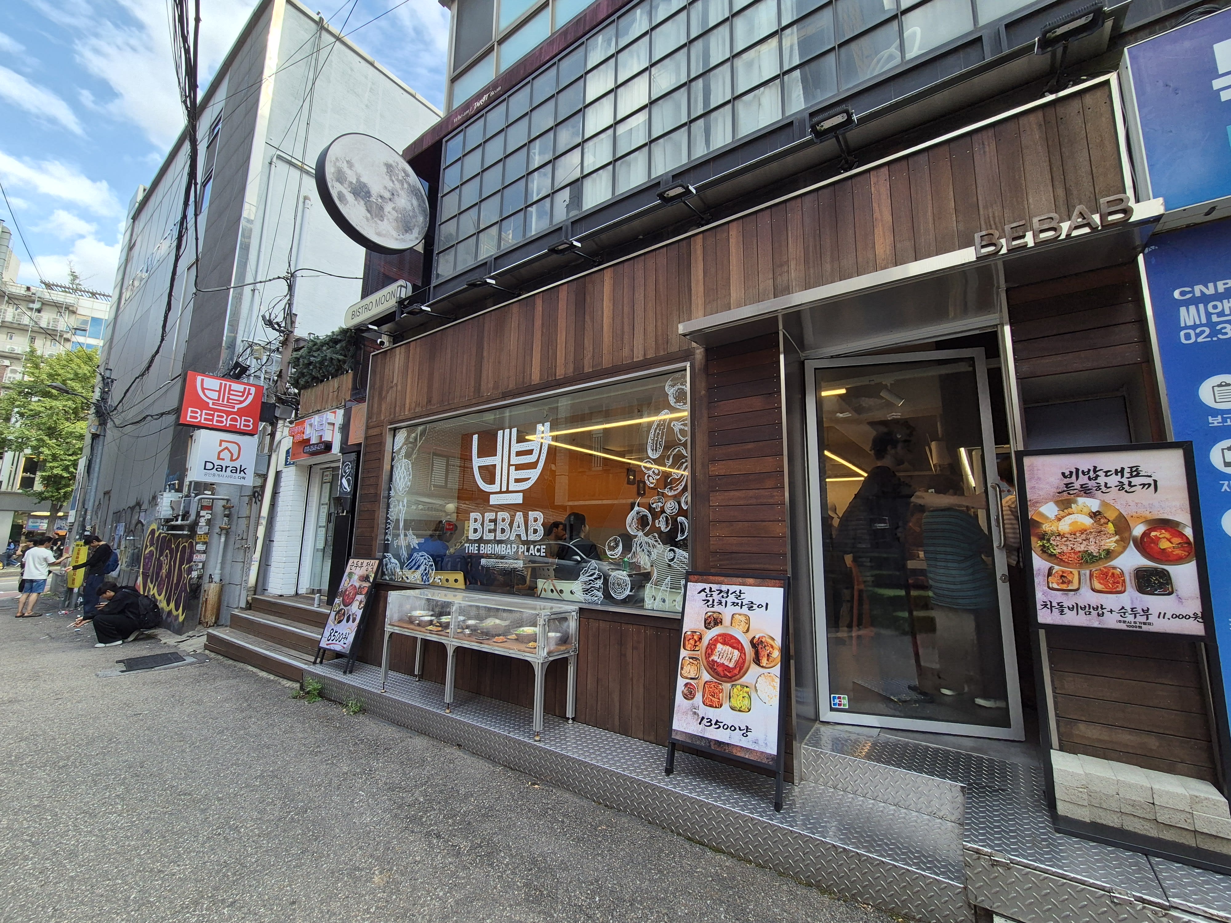 The BEBAB storefront with a window reading BEBAB THE BIBIMBAP PLACE, a round moon-shaped sign above, and menu stands out front