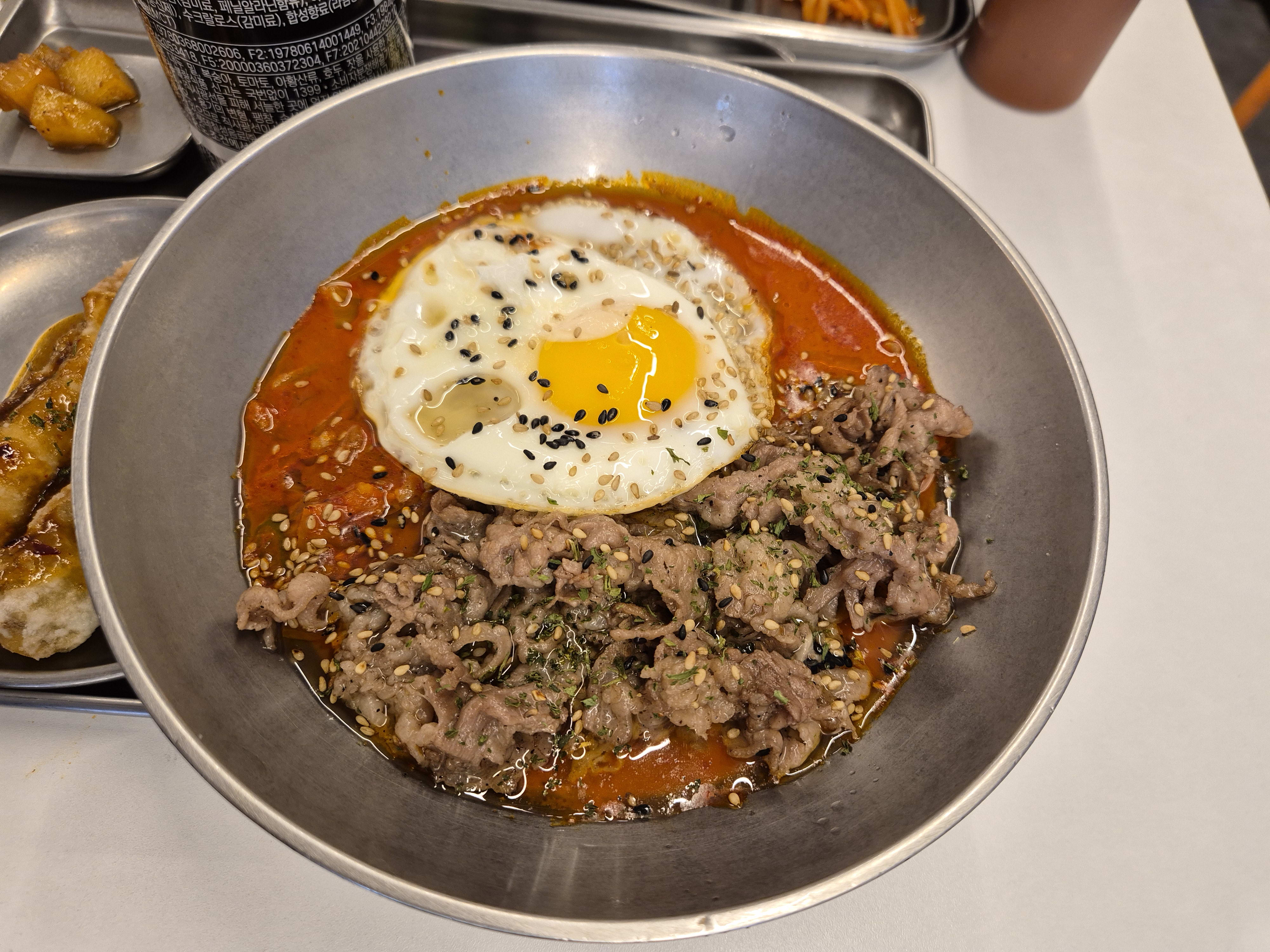 Chadol jjageuri in a metal bowl with a fried egg, sesame seeds and thin sliced beef in red broth