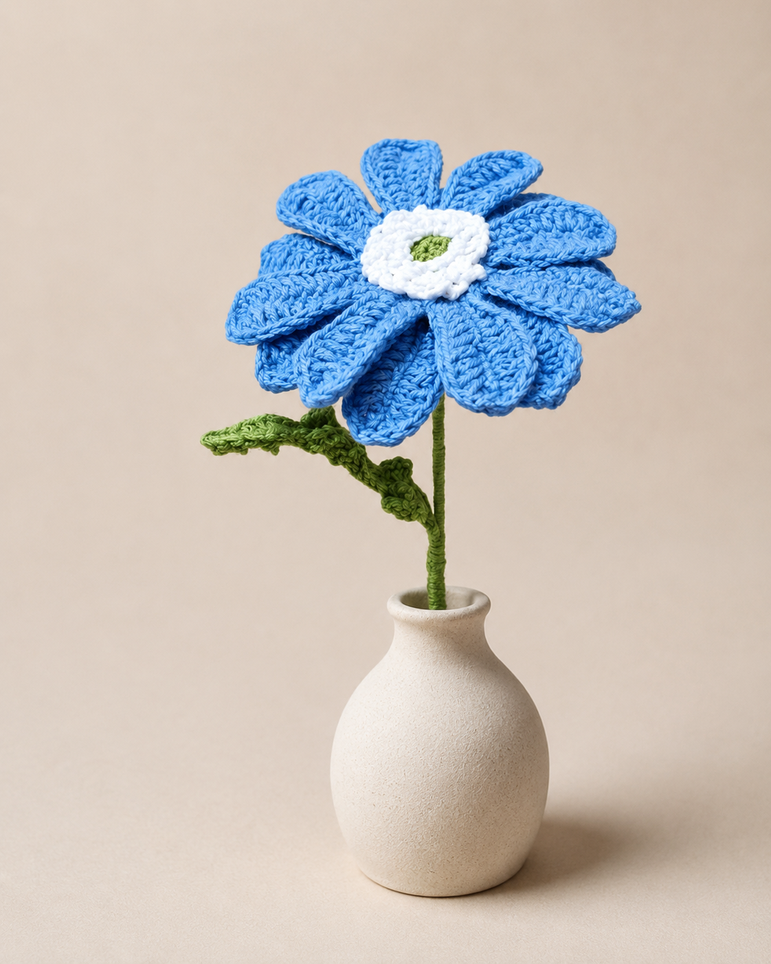 A single handcrafted royal blue crochet African daisy stem with a white center and green leaf, placed elegantly inside a small cream ceramic vase against a plain neutral beige backdrop.