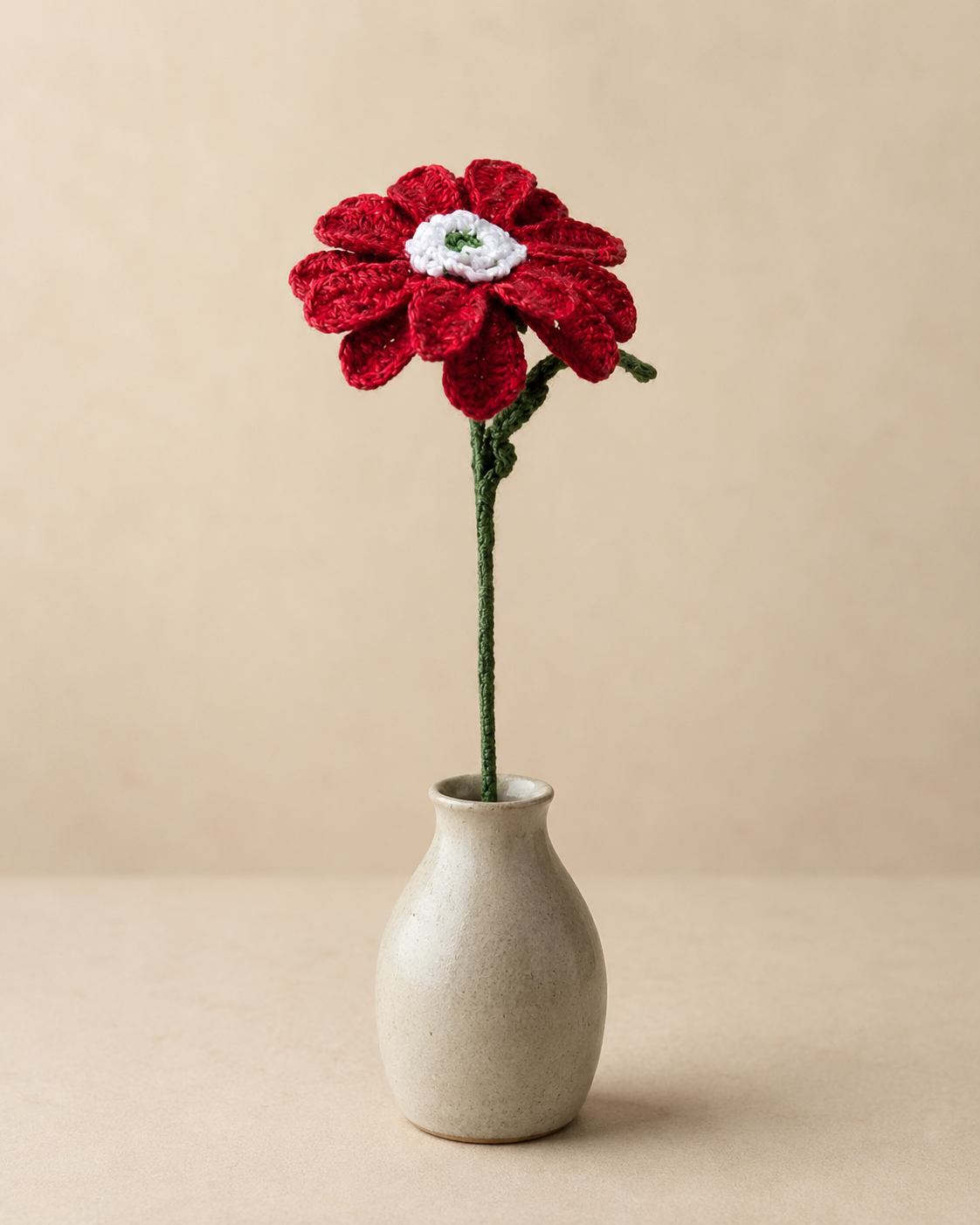 Crimson red African daisy crochet flower stem displayed in a small ceramic vase on a neutral background.