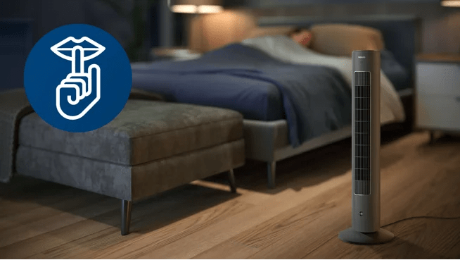 Ventilador de torre gris sobre el suelo de un dormitorio junto a la cama, mostrando su uso en el hogar