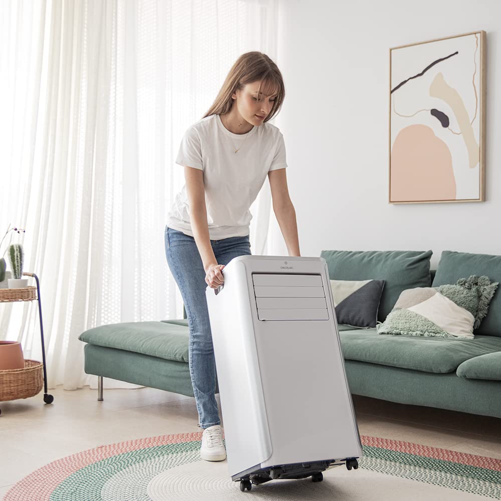 Mujer trasladando un aire acondicionado portátil Cecotec ForceClima 9100 blanco con ruedas en un salón moderno.