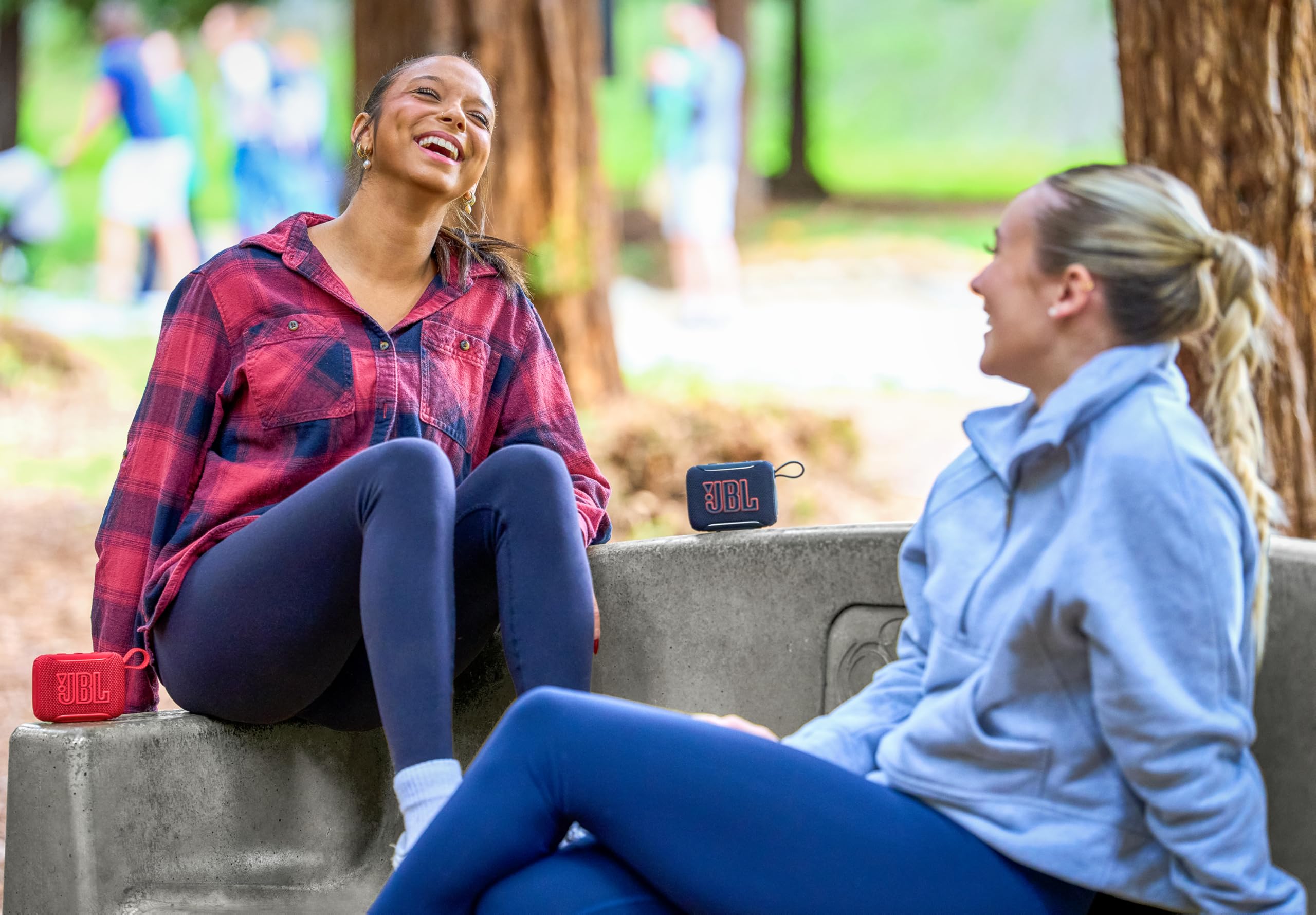 Dos amigas riendo en un parque con un altavoz JBL GO 5 Bluetooth portátil reproduciendo música entre ellas