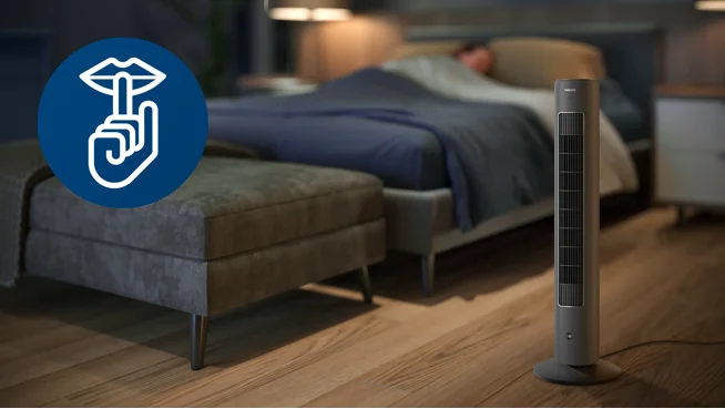 Ventilador de torre gris sobre el suelo de un dormitorio junto a la cama, mostrando su uso en el hogar