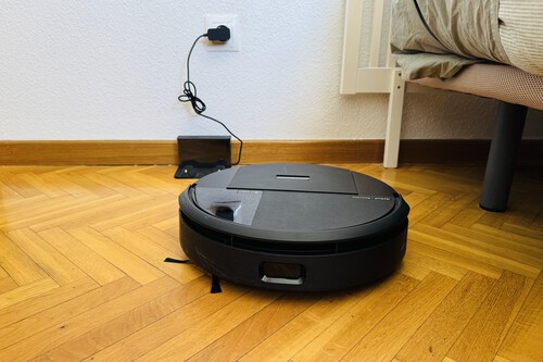 Robot aspirador negro en suelo de madera junto a su base de carga enchufada a la pared