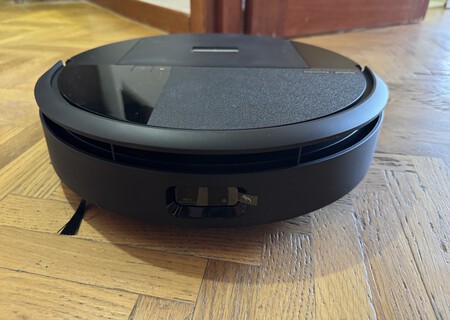 Robot aspirador negro redondo sobre suelo de madera, vista frontal mostrando sensores y contactos de carga