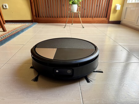 Robot aspirador negro circular limpiando suelo de baldosas en un salón con planta y barandilla de madera
