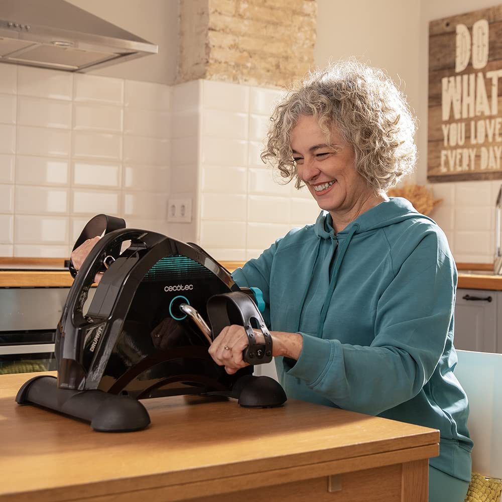 Mujer ejercitando brazos con pedales estáticos Cecotec DrumFit Cycle Compact sobre mesa de cocina.