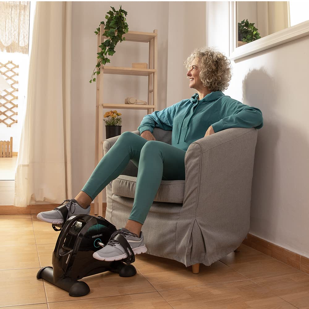 Mujer madura ejercitando piernas con pedales estáticos Cecotec DrumFit Cycle Compact en salón de casa.