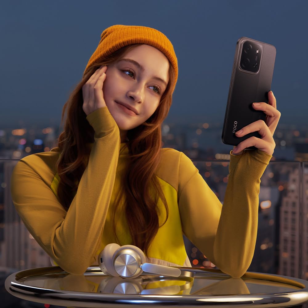 Young woman in yellow beanie holding black POCO C85 smartphone with dual 50MP camera, headphones on glass table, cityscape twilight background