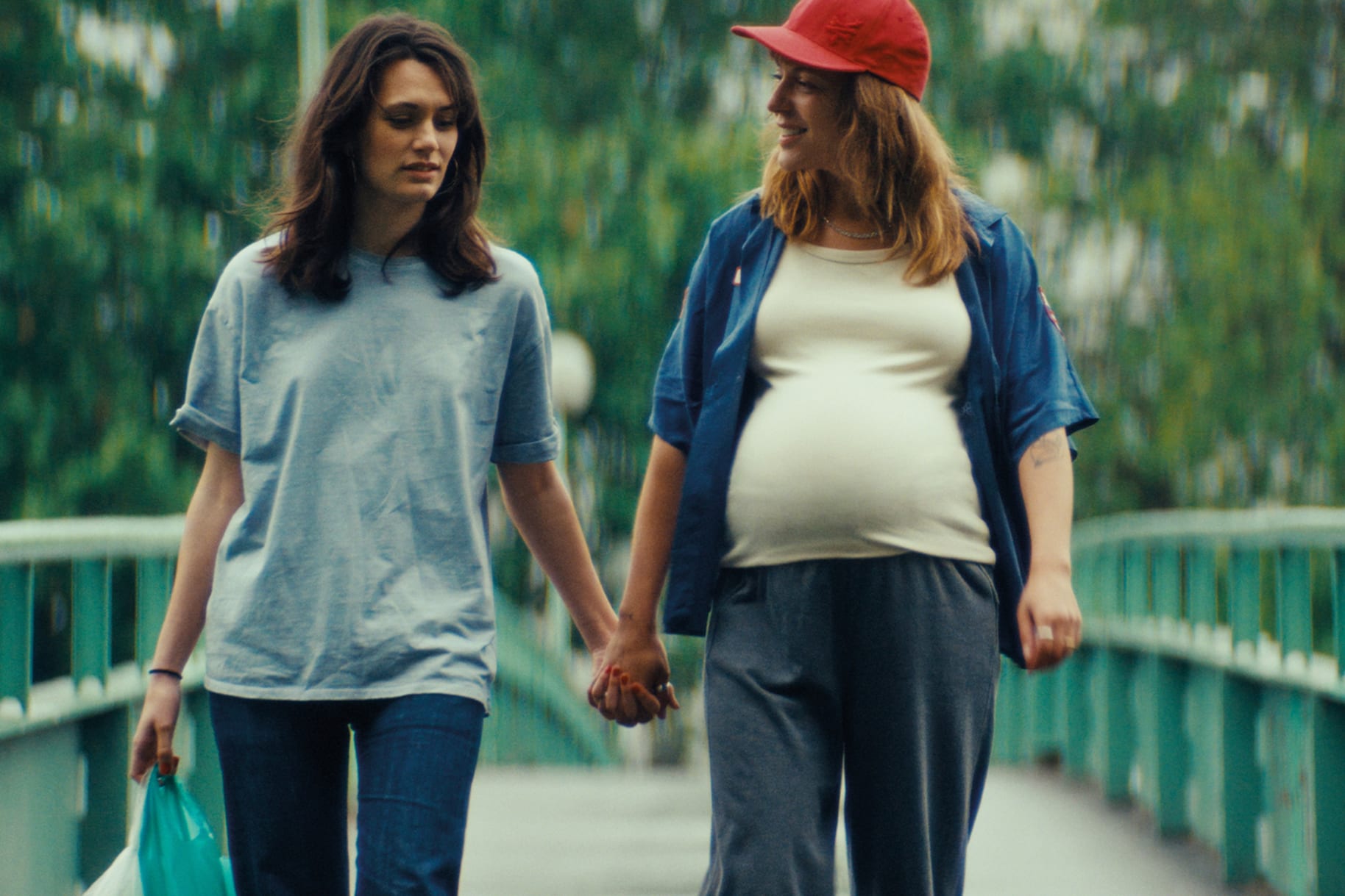 Zwei Frauen gehen Hand in Hand über eine Brücke, eine trägt eine rote Mütze und ein weisses T-Shirt und blaues Hemd, eine ein hellblaues T-Shirt annabelle