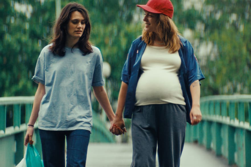 Zwei Frauen gehen Hand in Hand über eine Brücke, eine trägt eine rote Mütze und ein weisses T-Shirt und blaues Hemd, eine ein hellblaues T-Shirt annabelle