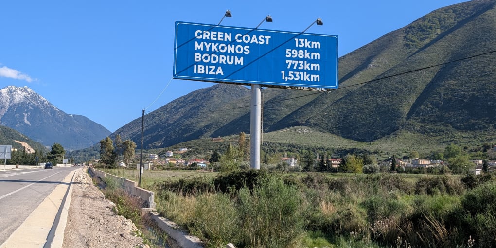 On the side of a highway in Orikum, Albania. A large sign says "Green Coast 13km, Mykonos 598km, Bodrum 773km, Ibiza 1531km."