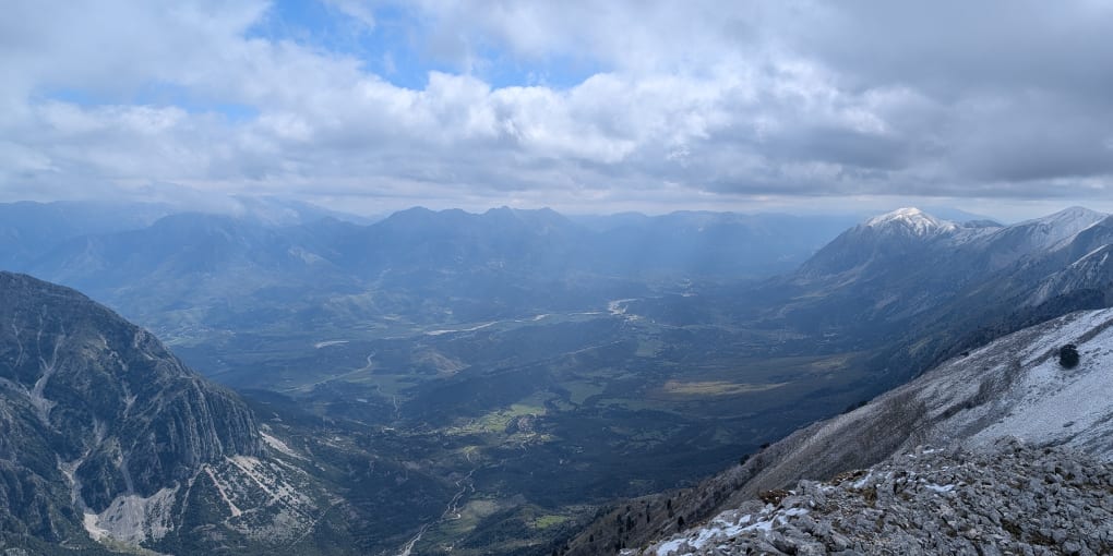 View looking Southeast from the Peak of Maja Paliskës. A large valley with light shining down between clouds.