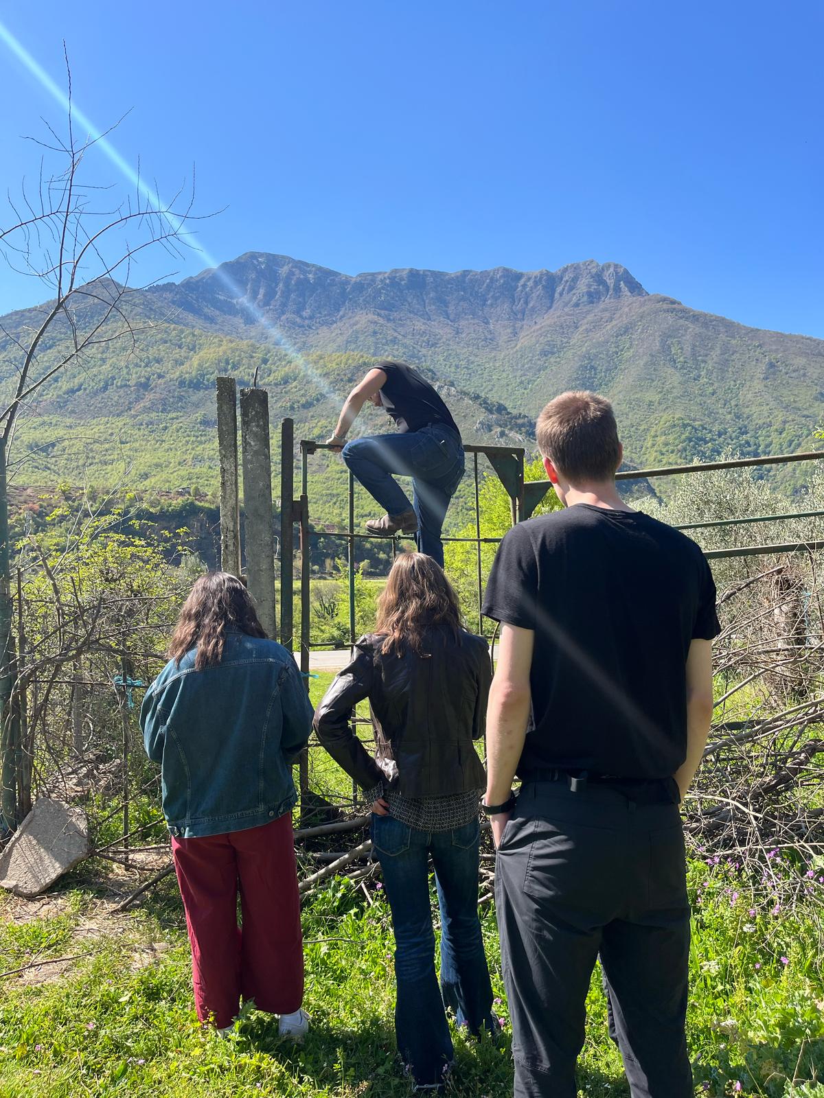 Three people watch someone climb a fence with high mountains in the background.