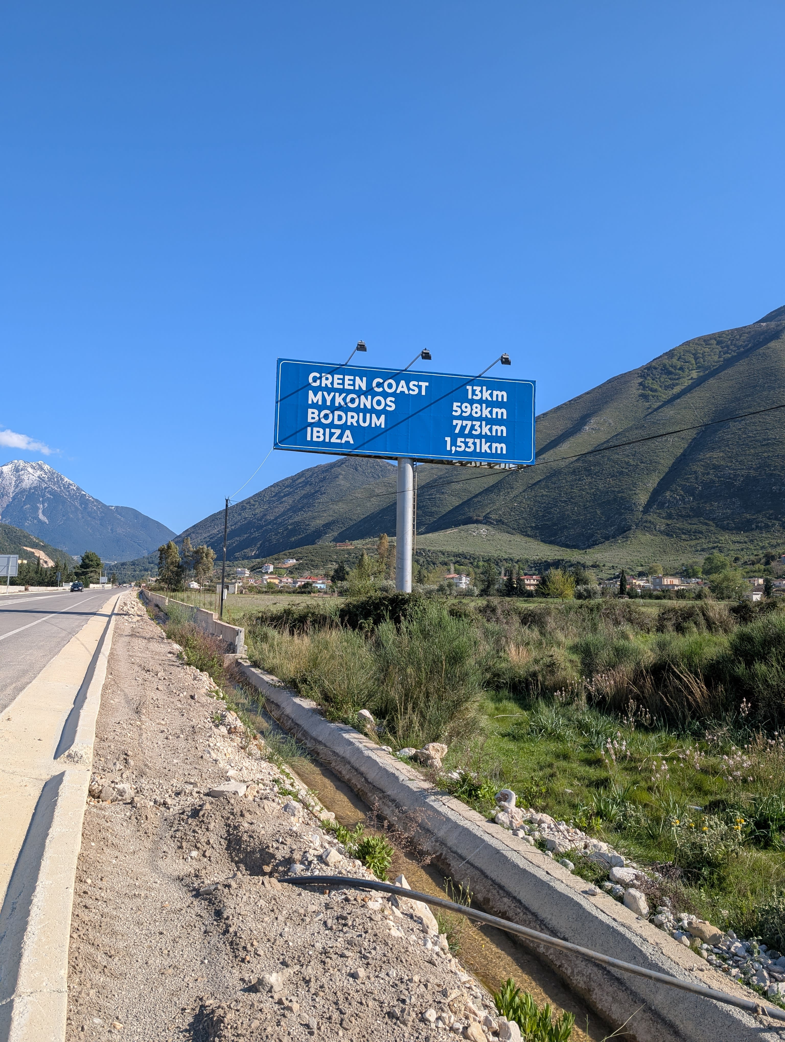 On the side of a highway in Orikum, Albania. A large sign says "Green Coast 13km, Mykonos 598km, Bodrum 773km, Ibiza 1531km."