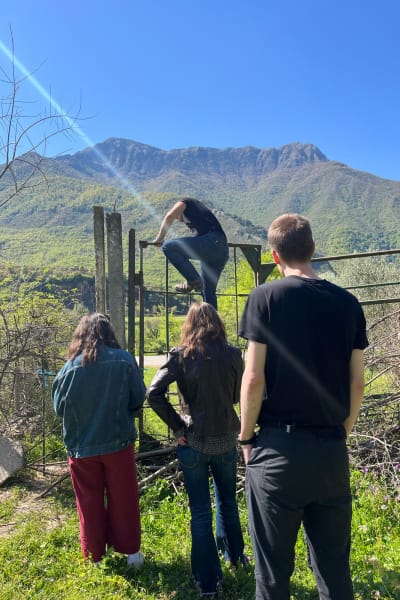 Three people watch someone climb a fence with high mountains in the background.