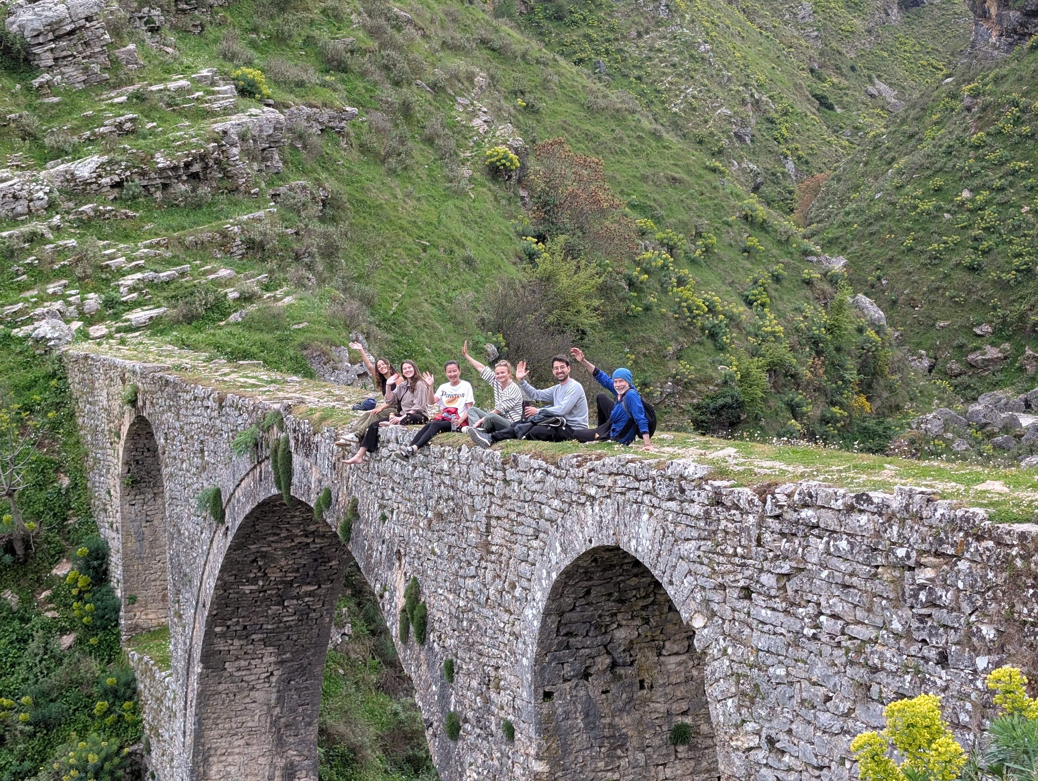 Six people are sitting on the Ali Pasha bridge which features stone arches. They wave to the camera.